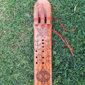 May include: A wooden musical instrument with intricate carvings and red leather straps. The instrument has a rectangular shape and is decorated with geometric patterns.
