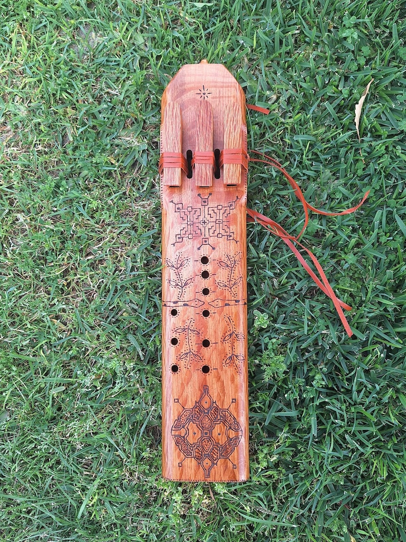 May include: A wooden musical instrument with intricate carvings and red leather straps. The instrument has a rectangular shape and is decorated with geometric patterns.
