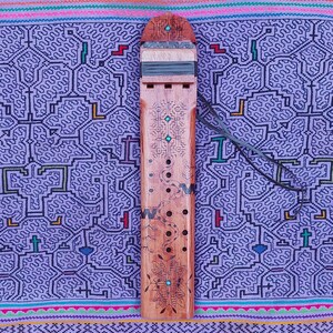 May include: A wooden flute with intricate carvings and turquoise accents. The flute is decorated with a snake design and has a series of holes along its length. It is resting on a purple and pink patterned fabric.