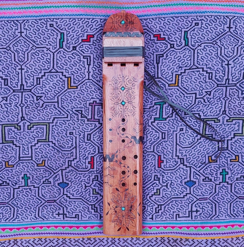 May include: A wooden flute with intricate carvings and turquoise accents. The flute is decorated with a snake design and has a series of holes along its length. It is resting on a purple and pink patterned fabric.