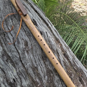 May include: A handcrafted wooden flute with a dark brown mouthpiece and a tan leather strap. The flute features decorative carvings and star-shaped accents along its length. The instrument rests on a weathered, textured wooden surface.