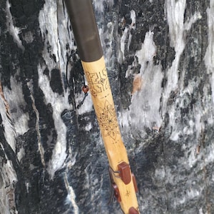May include: A handcrafted wooden flute with a light brown finish, featuring a dark brown leather wrap and decorative carvings. The flute has brown leather accents and is leaning against a textured tree trunk.