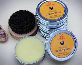 Può includere: Due barattoli di metallo aperti di balsamo per barba con un'etichetta gialla che recita "Heavenly Beard Balm, Net Wt. 1.88 oz, Made in California, USA". I barattoli sono su un tavolo accanto a una spazzola di legno e una paletta di legno piena di piccoli cristalli marroni.