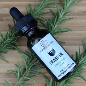 May include: A brown glass bottle with a dropper top filled with beard oil. The label is white with green text that reads "Pink Blossom Soap Company" and "Beard Oil Rosemary Mint". The label also includes a small clover graphic and a cartoon beard. The bottle is sitting on a wooden surface with green rosemary sprigs.