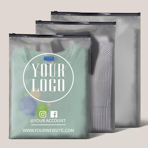 以下が含まれることがあります： 黒いジッパーが付いた透明なプラスチック製の衣類バッグが3つ。前面のバッグには、白い円形のロゴに「YOUR LOGO」と書かれた薄緑色のTシャツが入っています。ソーシャルメディアのアイコンとウェブサイトのアドレスも表示されています。