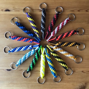 Puede incluir: Una colección de 15 llaveros coloridos hechos con paracord. Cada llavero tiene una combinación de colores diferente y está atado con un nudo en la parte superior. Los llaveros están dispuestos en círculo sobre una superficie de madera.