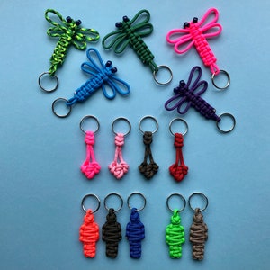 Könnte beinhalten: Sortiment an bunten Paracord-Schlüsselanhängern mit Libellen- und Menschenfiguren. Die Schlüsselanhänger sind aus verschiedenen farbigen Paracord gefertigt und haben Metallschlüsselringe.