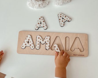 May include: A wooden puzzle with the name "AMAI" spelled out in individual letters. Each letter is decorated with a floral pattern. A child's hand is placing the letter "A" into the puzzle.