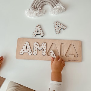 May include: A wooden puzzle with the name "AMAI" spelled out in individual letters. Each letter is decorated with a floral pattern. A child's hand is placing the letter "A" into the puzzle.