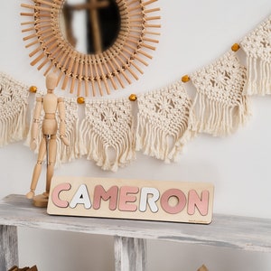 Op de afbeelding: Een houten naam puzzel met de naam "CAMERON" gespeld in roze en witte letters. De puzzel ligt op een wit rek met een houten pop en een macramee-slinger die erboven hangt.