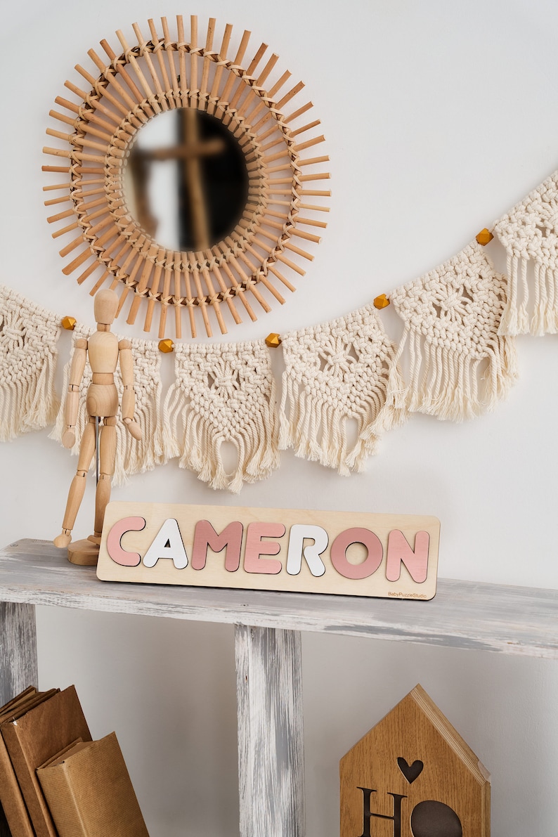 Op de afbeelding: Een houten naam puzzel met de naam "CAMERON" gespeld in roze en witte letters. De puzzel ligt op een wit rek met een houten pop en een macramee-slinger die erboven hangt.