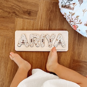 Peut inclure: Un puzzle en bois avec le nom "ARIYA" écrit en grandes lettres. Les lettres sont décorées d'un motif floral dans des tons de brun, beige et blanc. Le puzzle est sur un sol en bois et les pieds d'un enfant sont visibles en bas de l'image.