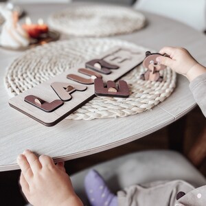 May include: A wooden puzzle with the name "LAURE" spelled out in brown letters on a light brown background. A small brown bear figurine is on the table next to the puzzle.