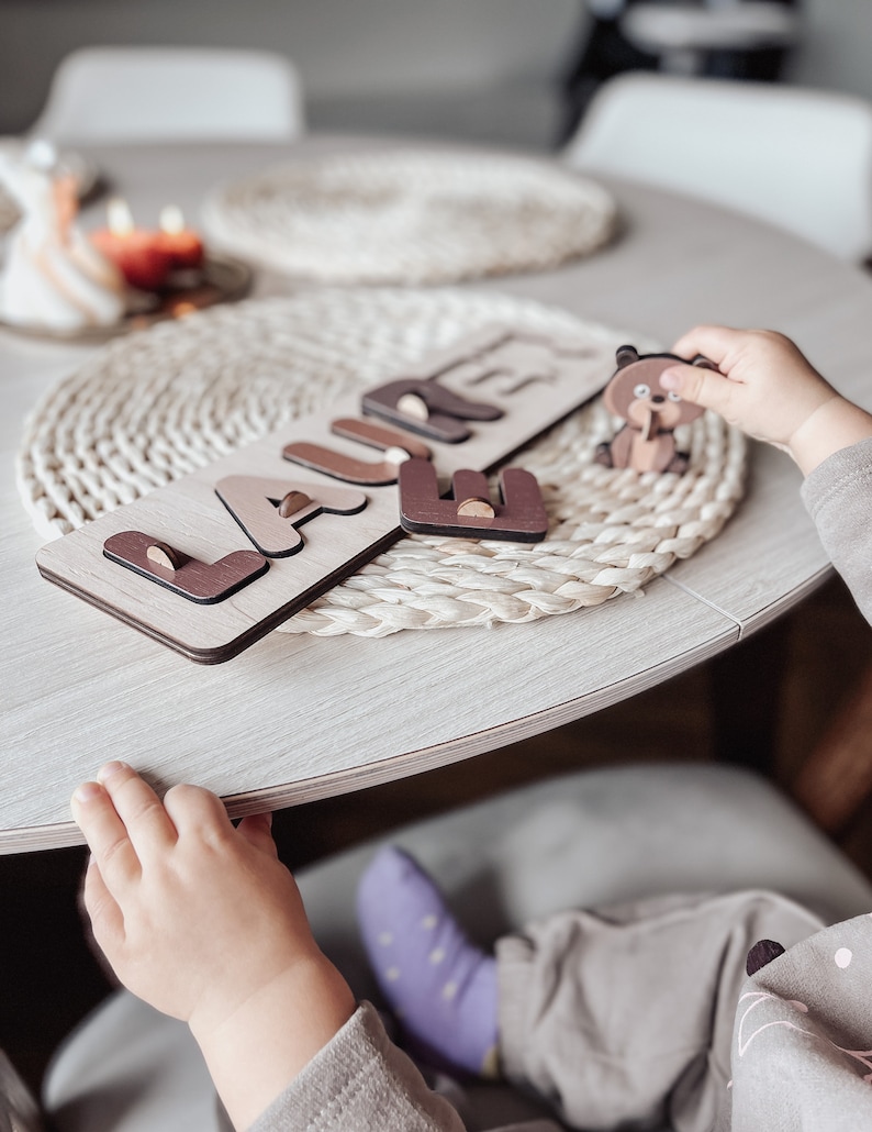 May include: A wooden puzzle with the name "LAURE" spelled out in brown letters on a light brown background. A small brown bear figurine is on the table next to the puzzle.