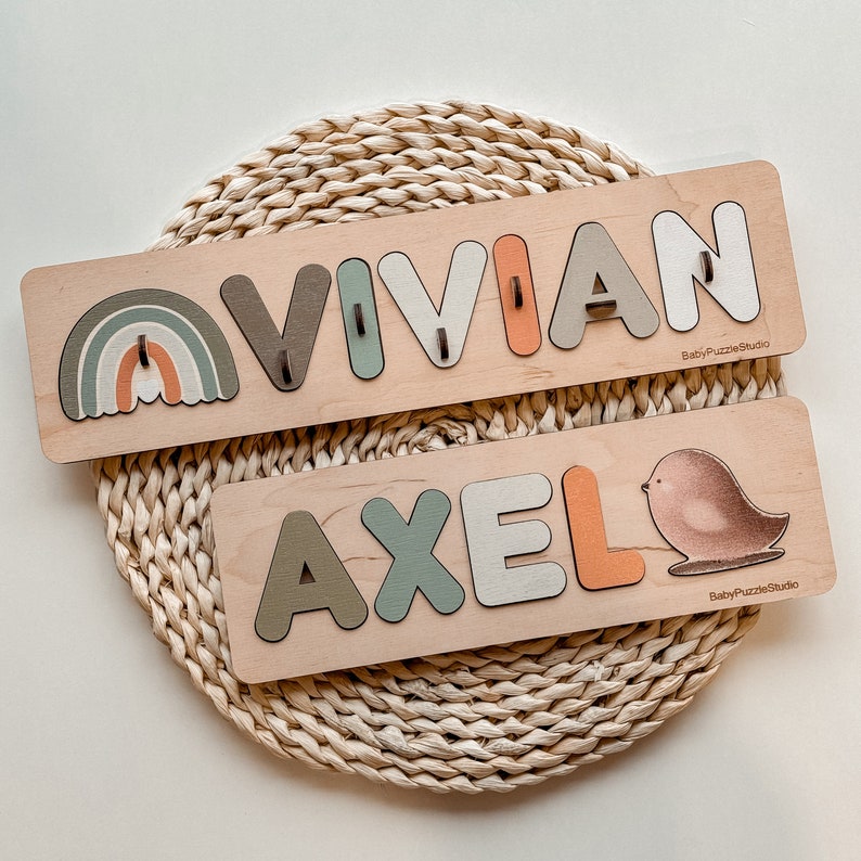 Op de afbeelding: Twee houten puzzelplanken met de namen "Vivian" en "Axel" gespeld in kleurrijke letters. De Vivian-plank heeft een regenboogontwerp en de Axel-plank heeft een klein vogelontwerp. Beide planken hebben de tekst "BabyPuzzleStudio" erop gedrukt.