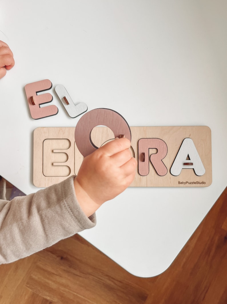 Op de afbeelding: Een houten puzzel met de naam "ELORA" gespeld in roze en witte letters. De letters worden door een kinderhand in de puzzel geplaatst. De puzzel ligt op een licht houten oppervlak. De tekst "BabyPuzzleStudio" staat in de rechterbenedenhoek van de puzzel.