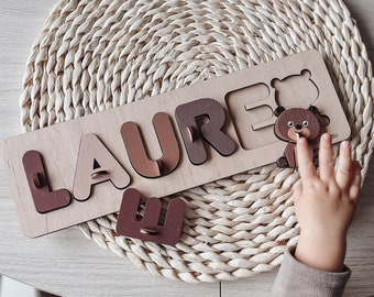 May include: Wooden puzzle with the name "LAURE" spelled out in brown letters. The puzzle pieces are shaped like the letters and fit into the corresponding slots. A small wooden bear is included as a puzzle piece.