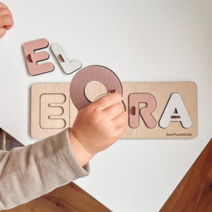 May include: A wooden puzzle with the name "ELORA" spelled out in pink and white letters. The letters are separate pieces that fit into the puzzle board. A child's hand is shown placing the letter "O" into the puzzle board. The puzzle board has the text "BabyPuzzleStudio" in the bottom right corner.
