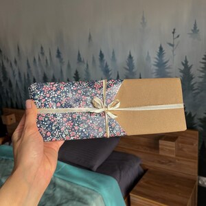 May include: A gift box wrapped in brown paper and a floral patterned paper with a white ribbon. The box is being held by a hand.