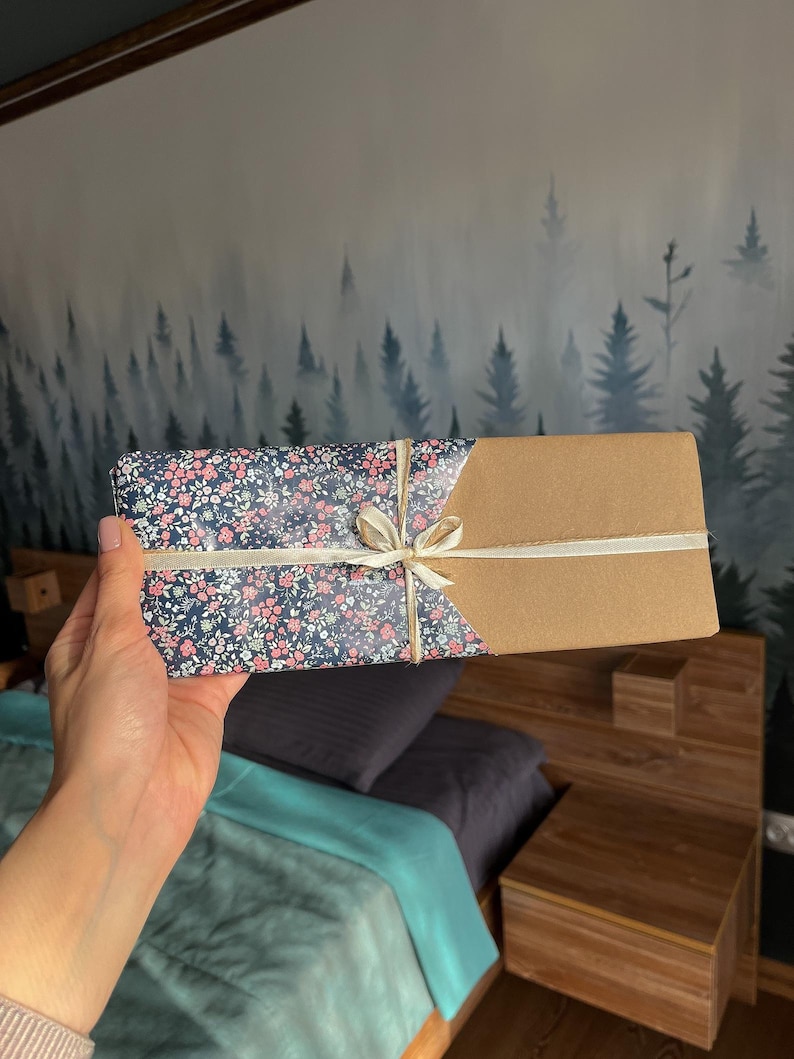 May include: A gift box wrapped in brown paper and a floral patterned paper with a white ribbon. The box is being held by a hand.