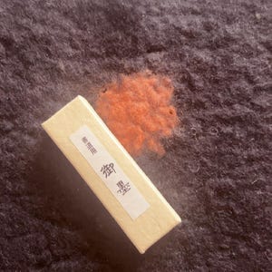 Könnte beinhalten: Eine rechteckige, hellfarbene Schachtel mit japanischem Text, wahrscheinlich für Kalligraphie-Tinte, liegt auf einer dunklen, strukturierten Oberfläche. Ein kleines, rotbraunes Tintenkissen befindet sich neben der Schachtel. Die Schachtel trägt japanischen Text.