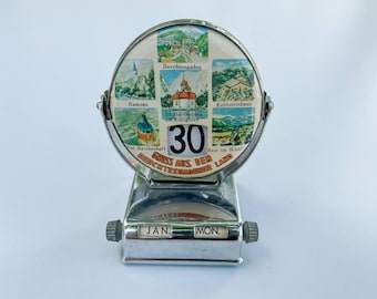 Pode incluir: Um calendário de mesa vintage em metal prateado. A face circular exibe imagens cênicas de Berchtesgaden, Alemanha, com a data "30" em destaque. O calendário mostra "JAN" e "MON" na parte inferior.