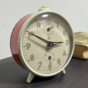 Könnte beinhalten: Vintage Peter Wecker in Rot und Weiß, mit cremefarbenem Zifferblatt und schwarzen Zeigern und Ziffern. Er hat ein kleines Zifferblatt für die Sekunden. Die Uhr steht auf einer dunklen Oberfläche, im Hintergrund ist ein Buch zu sehen.