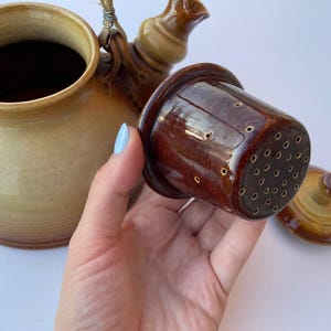 Puede incluir: Una tetera de cerámica con un infusor y tapa marrones. La tetera es de color beige claro con detalles marrones. El infusor es de color marrón oscuro con muchos agujeros pequeños. La tapa es de color marrón a juego.
