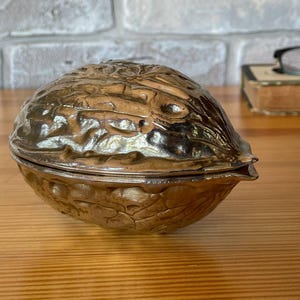 Puede incluir: Un recipiente decorativo en forma de nuez, de color plateado. El recipiente es de metal y tiene una superficie texturizada y detallada. La mitad superior de la nuez se levanta para revelar el interior. El recipiente está sobre una superficie de madera.