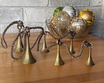 May include: A decorative arrangement featuring brass-colored bell-shaped ornaments connected by a brown rope. A glass bowl filled with colorful Christmas ornaments sits in the background. The ornaments are gold, white, green, and yellow.