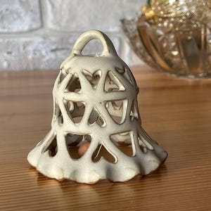 Puede incluir: Adorno en forma de campana de cerámica blanquecina con asa de bucle. La campana presenta un diseño de celosía con recortes en forma de corazón. La base tiene un borde con volantes. El adorno está sobre una superficie de madera.