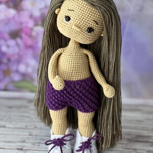 May include: A crocheted doll with long brown hair, wearing a purple crocheted skirt and white and purple crocheted shoes.