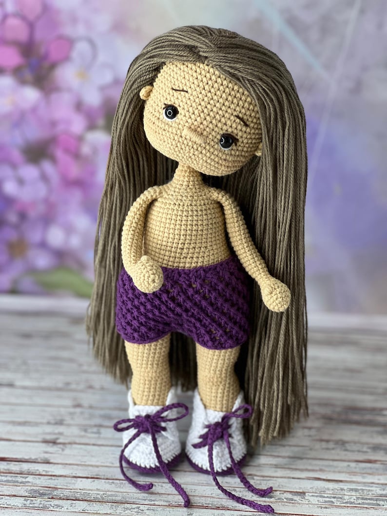 May include: A crocheted doll with long brown hair, wearing a purple crocheted skirt and white and purple crocheted shoes.