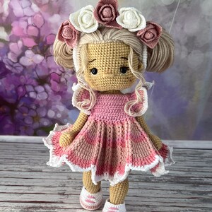 May include: A crocheted doll wearing a pink dress with white trim and a flower crown. The doll has blonde hair and brown eyes.