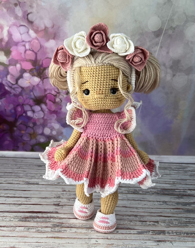 May include: A crocheted doll wearing a pink dress with white trim and a flower crown. The doll has blonde hair and brown eyes.