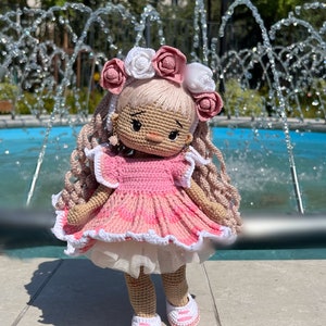 May include: A crocheted doll wearing a pink dress with white trim and a white and pink flower crown. The doll has blonde hair and brown eyes. The doll is standing in front of a fountain.