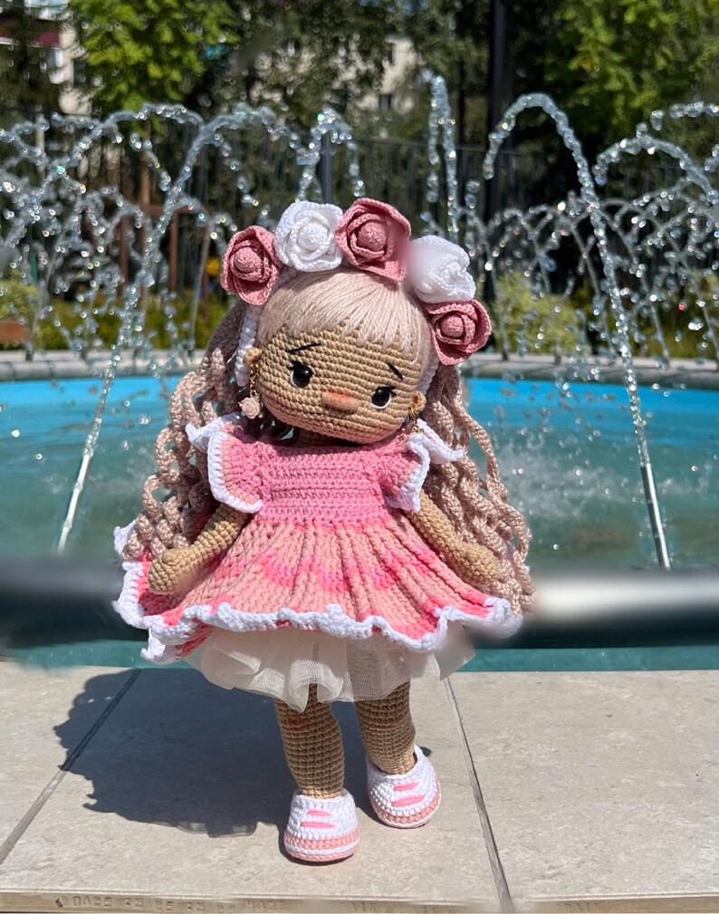 May include: A crocheted doll wearing a pink dress with white trim and a white and pink flower crown. The doll has blonde hair and brown eyes. The doll is standing in front of a fountain.