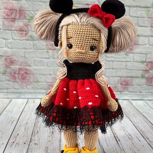 May include: A crocheted doll with blonde hair, wearing a red and black polka dot dress with a black lace trim, black Mickey Mouse ears with a red bow, and yellow shoes.