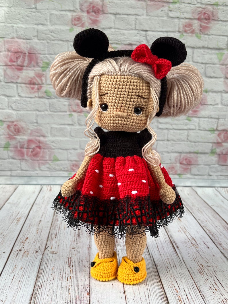 May include: A crocheted doll with blonde hair, wearing a red and black polka dot dress with a black lace trim, black Mickey Mouse ears with a red bow, and yellow shoes.