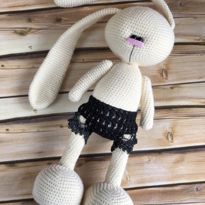 Puede incluir: Un conejito blanco de crochet con orejas largas y un borde de encaje negro en la parte inferior. El conejito est&aacute; acostado sobre una superficie de madera.