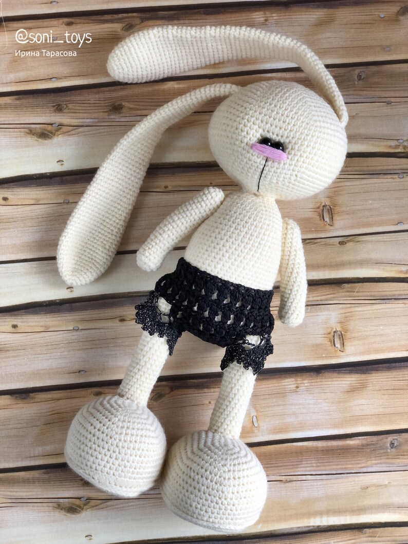 Puede incluir: Un conejito blanco de crochet con orejas largas y un borde de encaje negro en la parte inferior. El conejito est&aacute; acostado sobre una superficie de madera.