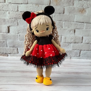 May include: A crocheted doll with blonde hair, wearing a red and black polka dot dress with black lace trim and black mouse ears. The doll is wearing yellow shoes.