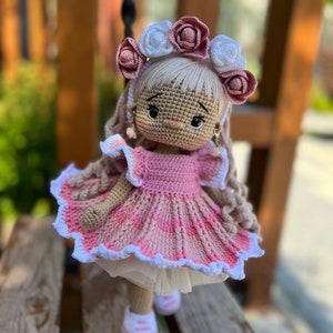 May include: A crocheted doll wearing a pink dress with white lace trim and a flower crown. The doll has long blonde hair and brown eyes.