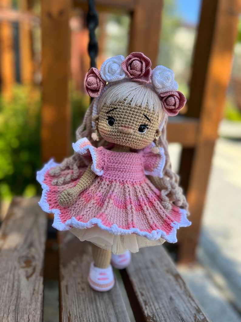 May include: A crocheted doll wearing a pink dress with white lace trim and a flower crown. The doll has long blonde hair and brown eyes.