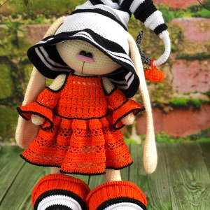 Puede incluir: Un conejito de crochet con un sombrero a rayas blanco y negro y un vestido naranja brillante. El conejito lleva zapatos a rayas blanco y negro. El conejito est&aacute; de pie sobre una superficie de madera con un fondo verde.