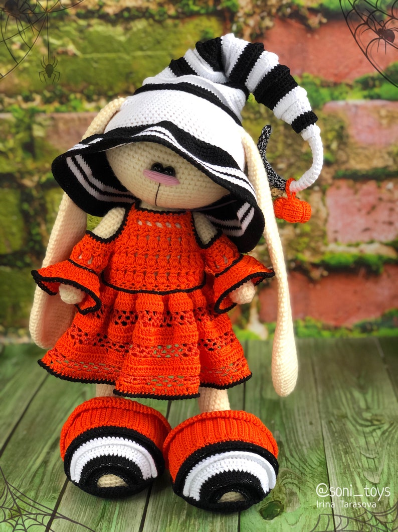 Puede incluir: Un conejito de crochet con un sombrero a rayas blanco y negro y un vestido naranja brillante. El conejito lleva zapatos a rayas blanco y negro. El conejito est&aacute; de pie sobre una superficie de madera con un fondo verde.