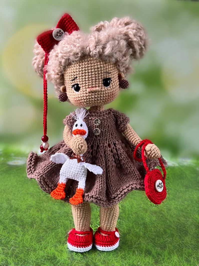 May include: A crocheted doll with brown hair, wearing a brown dress, red shoes, and a red bow. The doll is holding a white crocheted goose and a red purse.