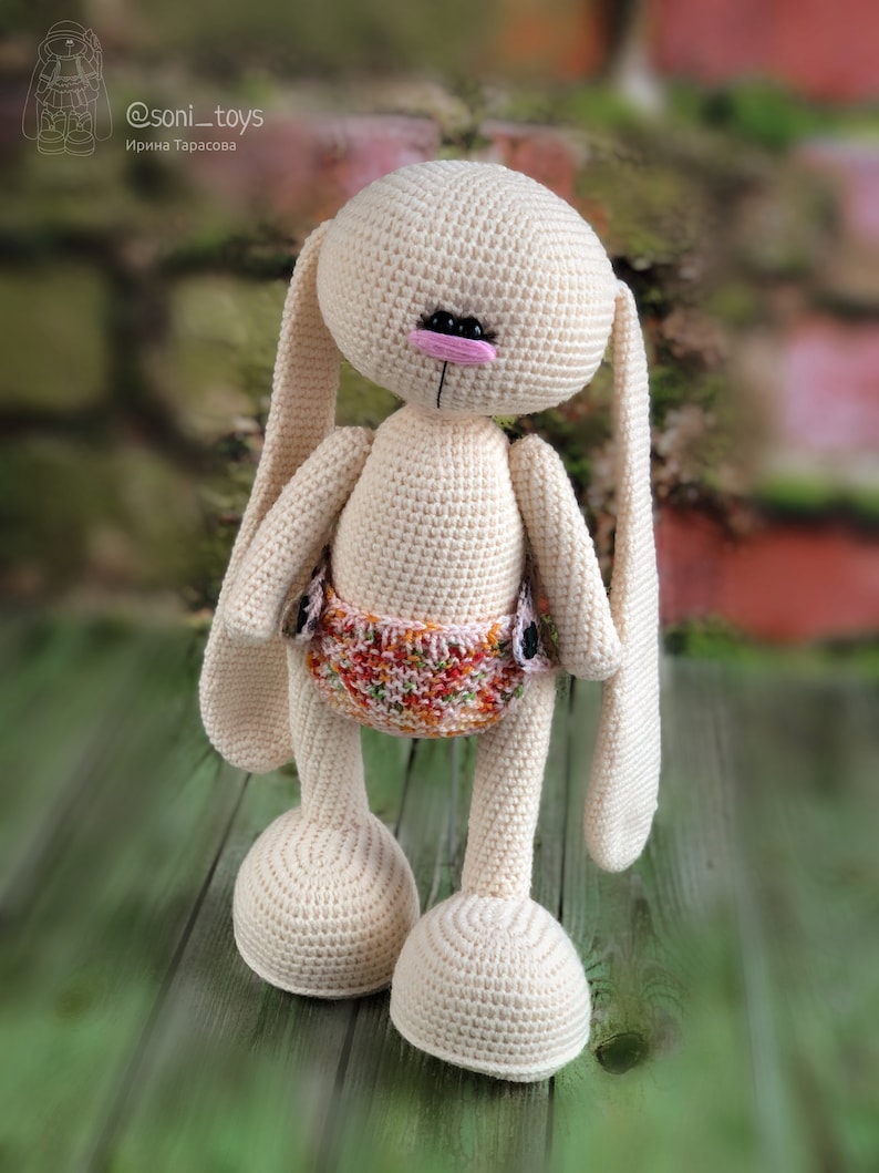 K&ouml;nnte beinhalten: Ein geh&auml;keltes Hasenspielzeug mit langen Ohren, einer rosa Nase und schwarzen Augen. Der Hase tr&auml;gt eine bunte Windel und steht auf einer Holzoberfl&auml;che. @soni_toys Irina Tarasova