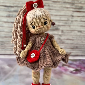 May include: A crocheted doll with brown hair, a red bow, and a brown dress. The doll is wearing red shoes and carrying a red purse with a button.