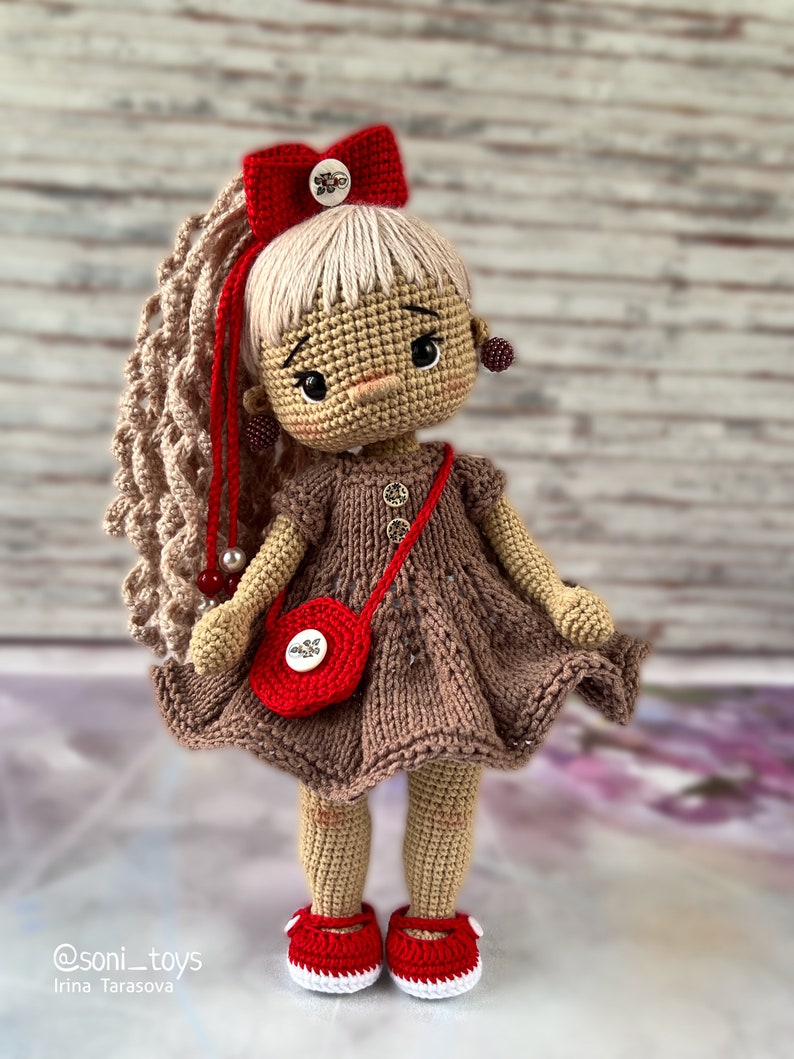 May include: A crocheted doll with brown hair, a red bow, and a brown dress. The doll is wearing red shoes and carrying a red purse with a button.